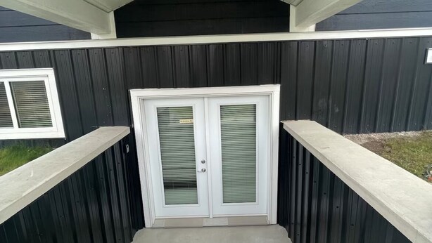 Un conjunto de escalones de hormigón que conducen a una puerta con panel de vidrio, flanqueada por paredes de madera negra y un techo blanco.