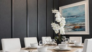 A dining table set with white chairs, plates, bowls, and wine glasses, with a white orchid in a black vase, against a dark wall with a framed painting.
