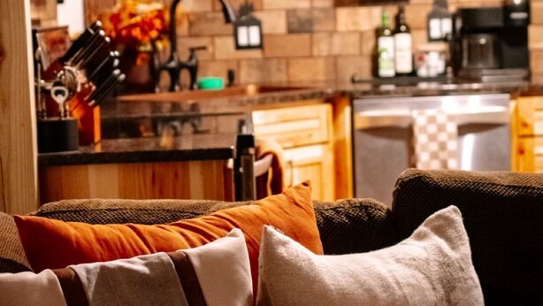 A cozy living room with a sofa and pillows, a kitchen in the background with wooden cabinets and a tiled backsplash, and a hanging light fixture. - Cozy 3-bedroom cabin in charming North East (North East)
