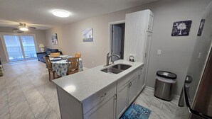 Een kleine keuken met een spoelbak, kastjes en een koelkast. Er is een eethoek met een tafel en stoelen, en uitzicht op de omgeving via een schuifdeur.