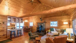 A log cabin interior with a stone fireplace, wooden furniture, and a kitchen area.