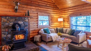 A cozy log cabin interior with a stone fireplace, a sofa set, and a view of the outdoors through a window.