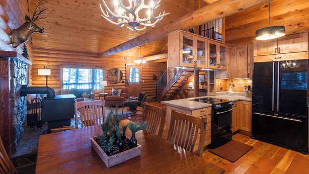 A log cabin interior with a dining area, kitchen, and a staircase leading to an upper level.