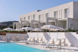 Un hôtel moderne doté d’un espace piscine comprenant des chaises longues et des parasols, d’une façade blanche et offrant une vue sur des collines en arrière-plan.