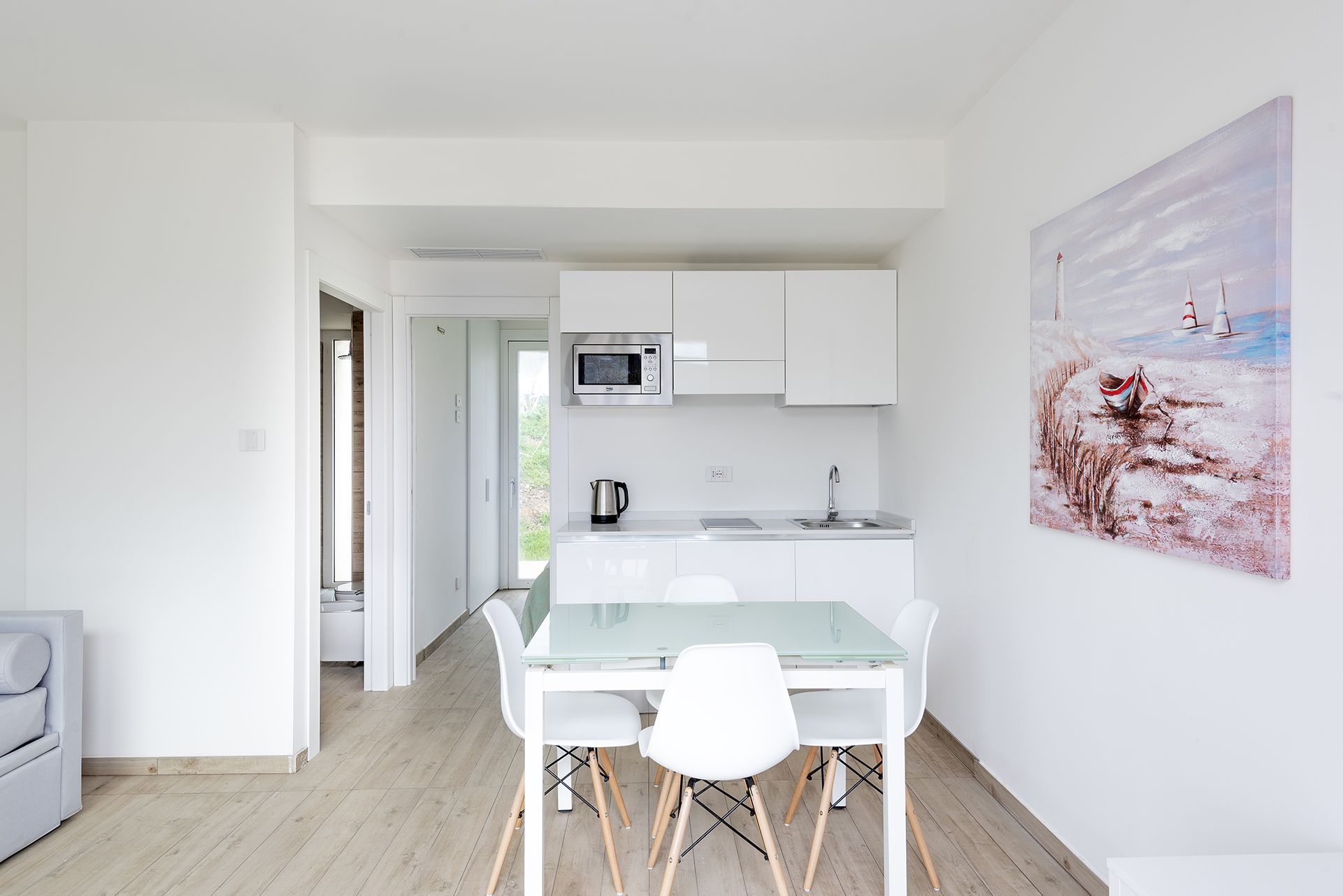 A modern kitchen with white cabinetry, a microwave, and a sink. There is a glass dining table with white chairs. A painting of a sailboat is on the wall.