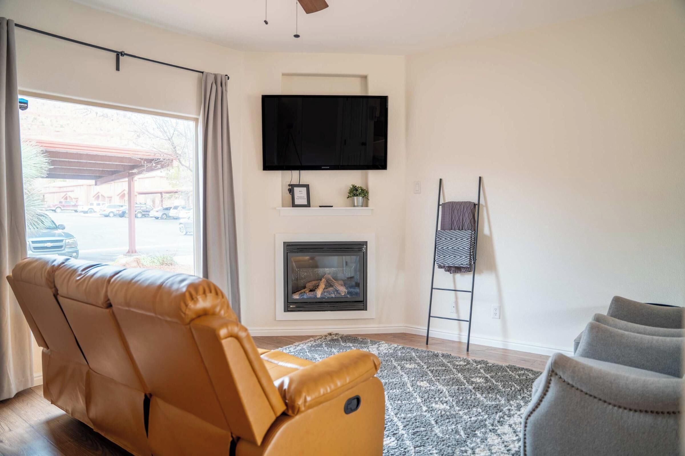 A living room with a leather sofa, a fireplace, a television mounted above it, and a view through a large window.