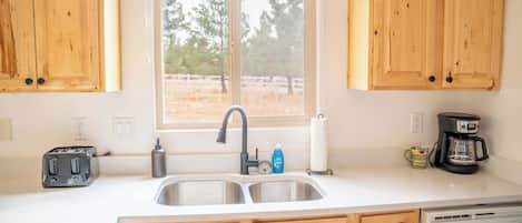 A kitchen with wooden cabinets, a double sink, a toaster, a coffee maker, and a window with a view of trees.
