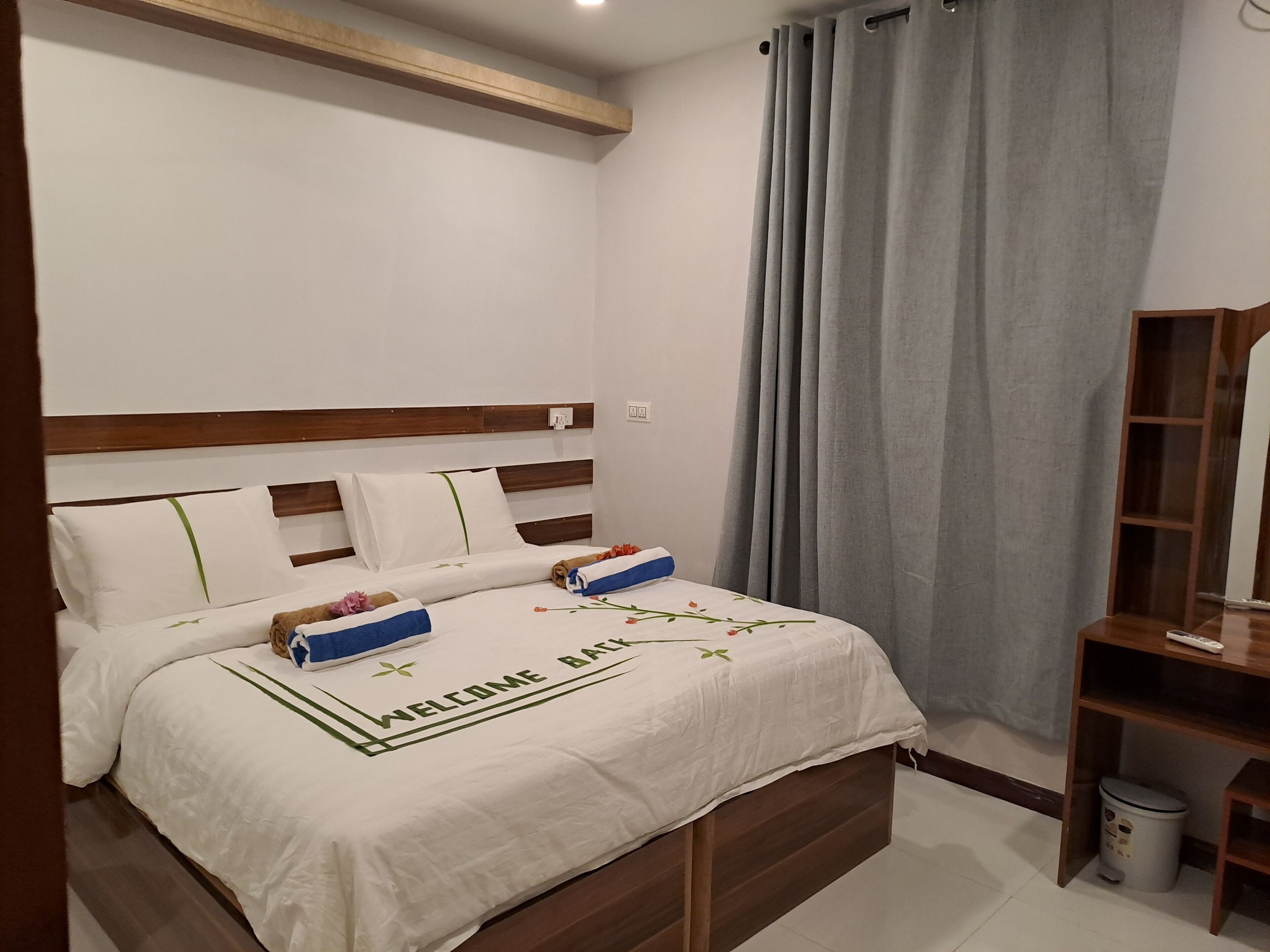 A neatly made bed with a white bedspread and pillows, a wooden headboard, and a grey curtain.