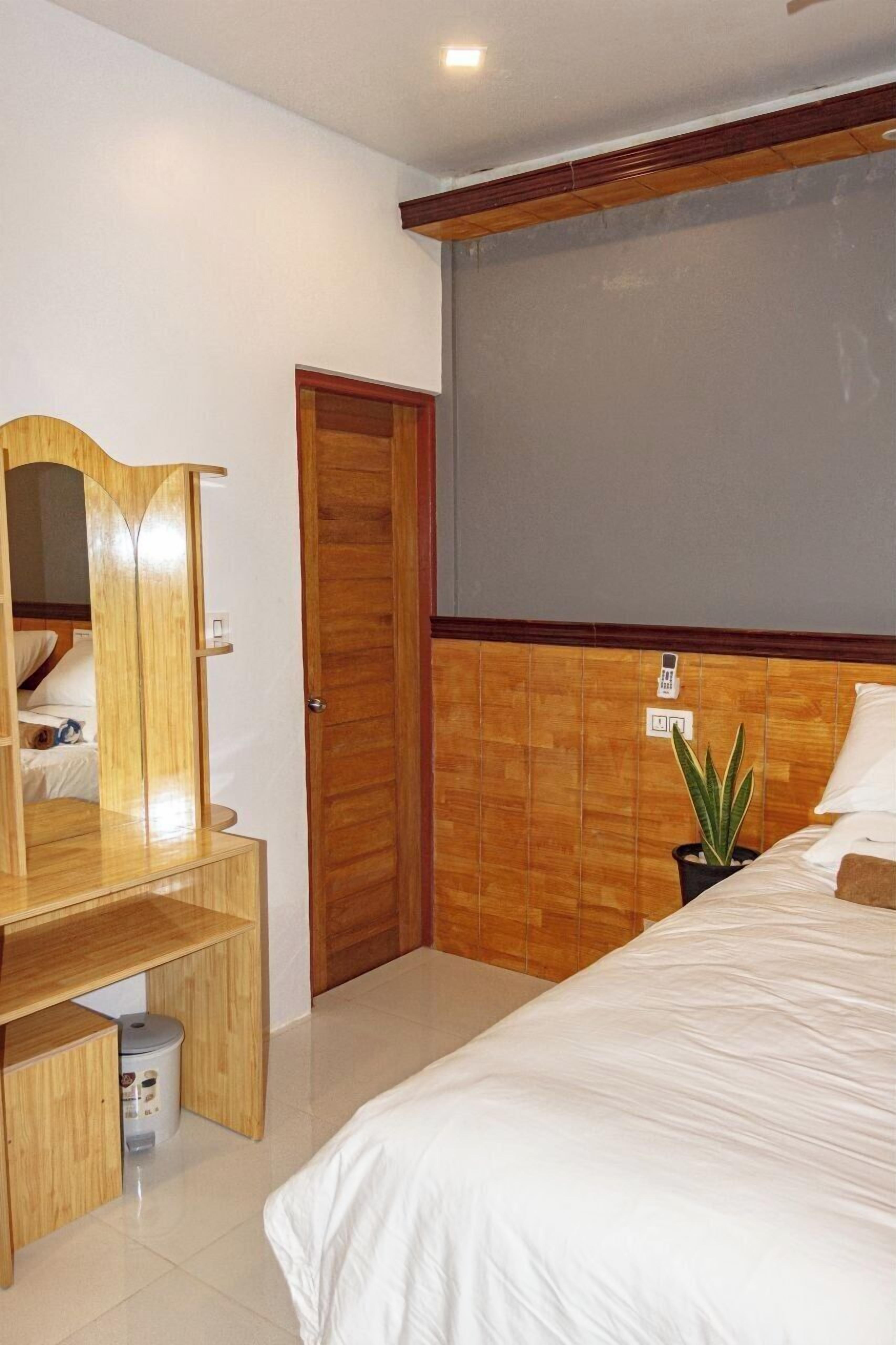 A hotel room with a wooden wardrobe, a bed, a small desk, a potted plant, and a wall-mounted phone.