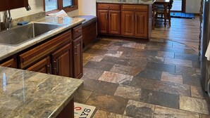 A kitchen with granite countertops, wooden cabinets, a stone tile floor, and a dining area with a wooden table and chairs. - Charming Lake House! (Wild Rose)
