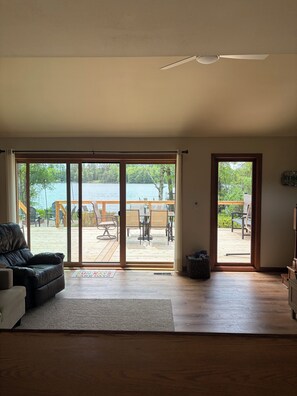 A room with a large sliding glass door leading to an outdoor deck with a view of a lake, a ceiling fan, and a black leather couch. - Charming Lake House! (Wild Rose)
