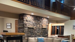A living room with a stone accent wall, a sectional sofa, a fireplace, and a dining area with a view of the balcony. - Charming Lake House! (Wild Rose)