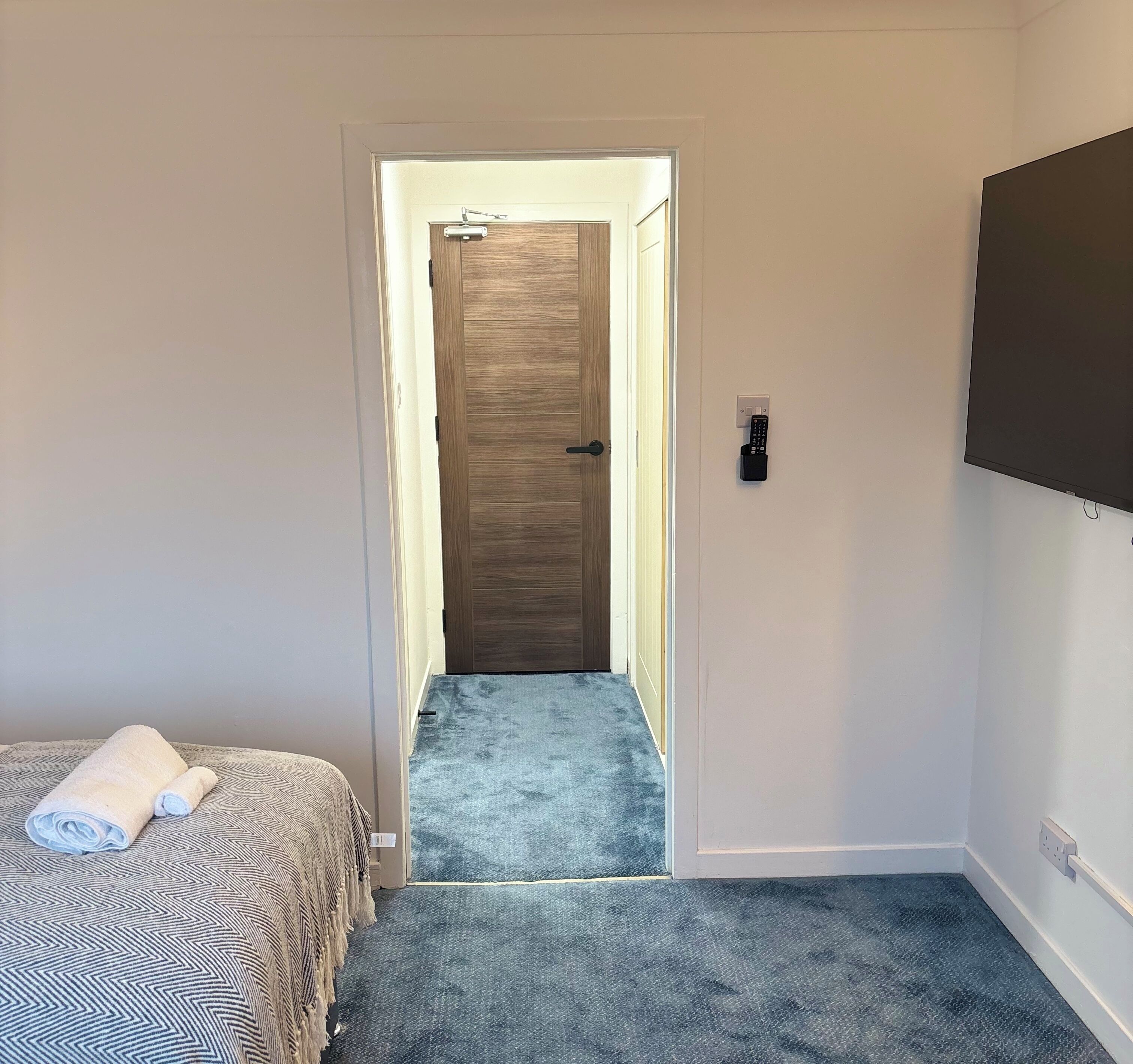 Ett hotellrum med en säng, en tv som är monterad på väggen och en dörr som leder ut till en korridor.