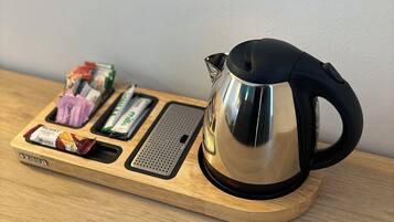 Una tetera de acero inoxidable sobre una bandeja de madera con una base calada y un pequeño compartimento para bocadillos.