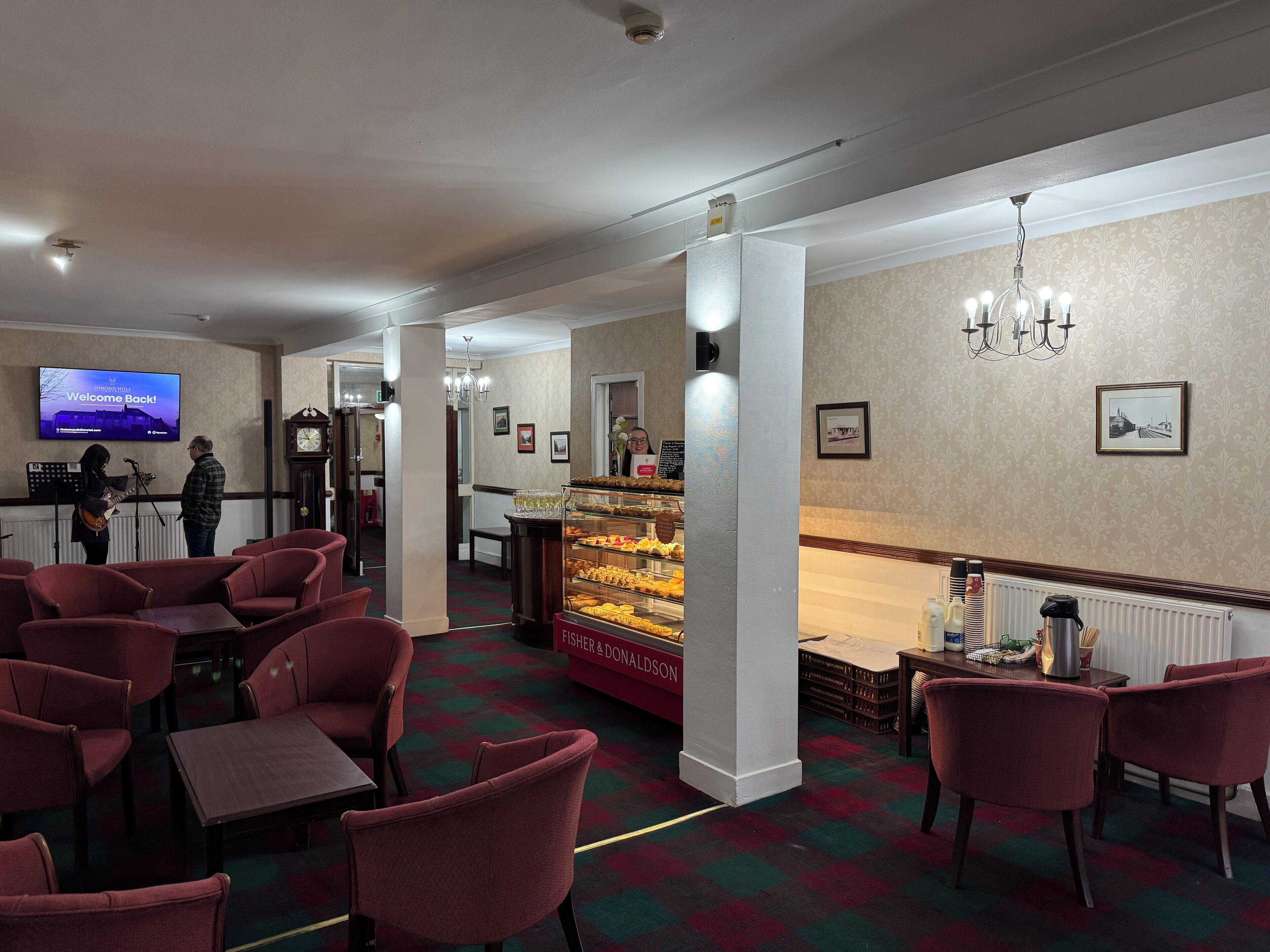 A hotel lobby with red chairs, a buffet counter, a television displaying 'Welcome Back!', and a chandelier.