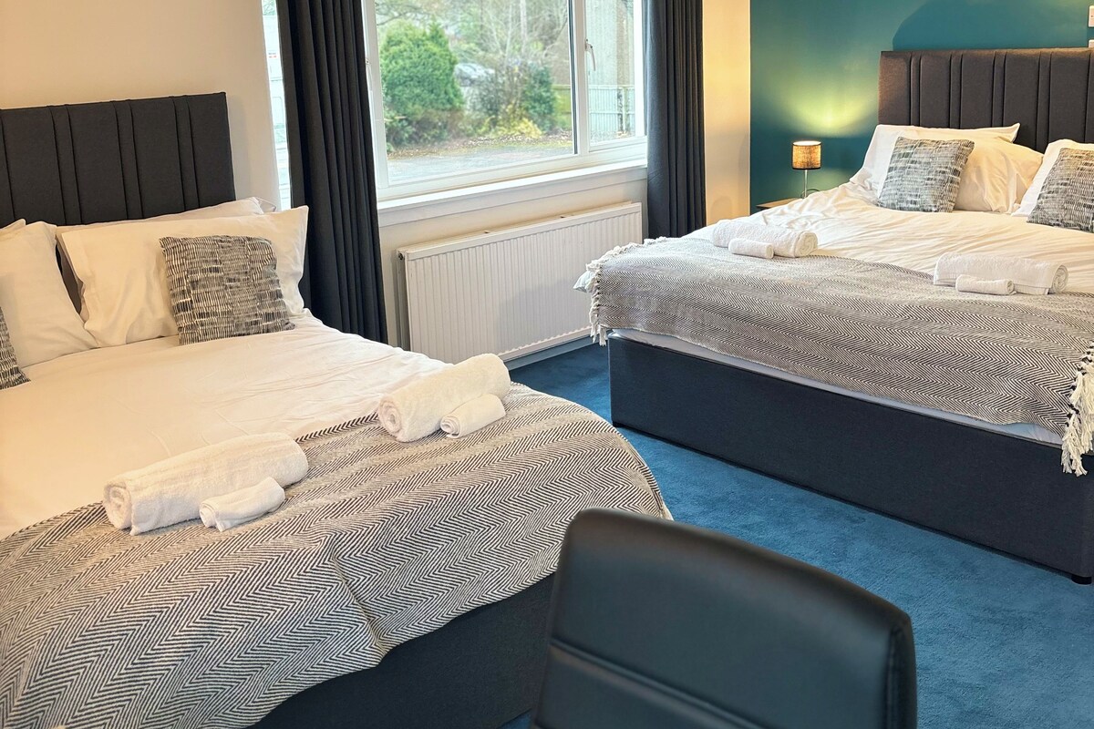 A hotel room with two beds, a view of greenery outside, and a blue carpet.