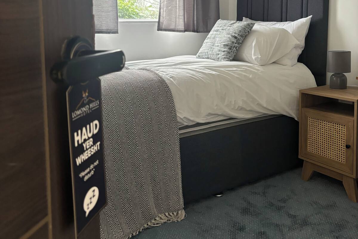 Ein Hotelzimmer mit Bett, Nachttisch, Lampe und einem Fenster mit Vorhängen.