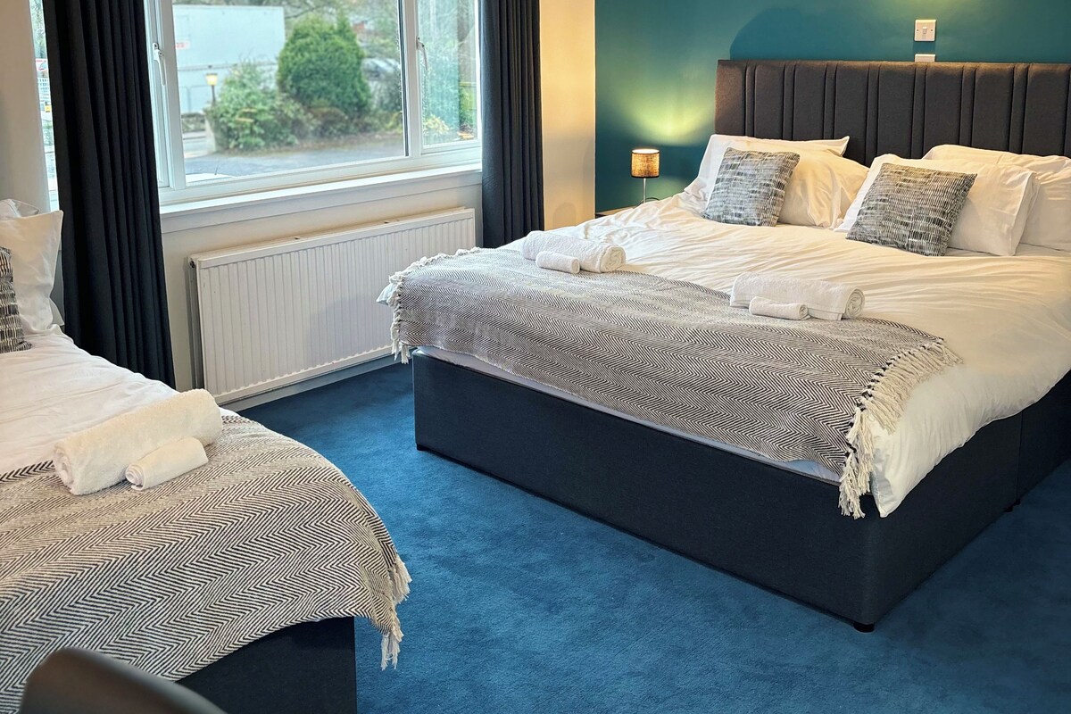 A hotel room with a large bed, a smaller bed, a window with curtains, a wall-mounted light, and a view of the outside.