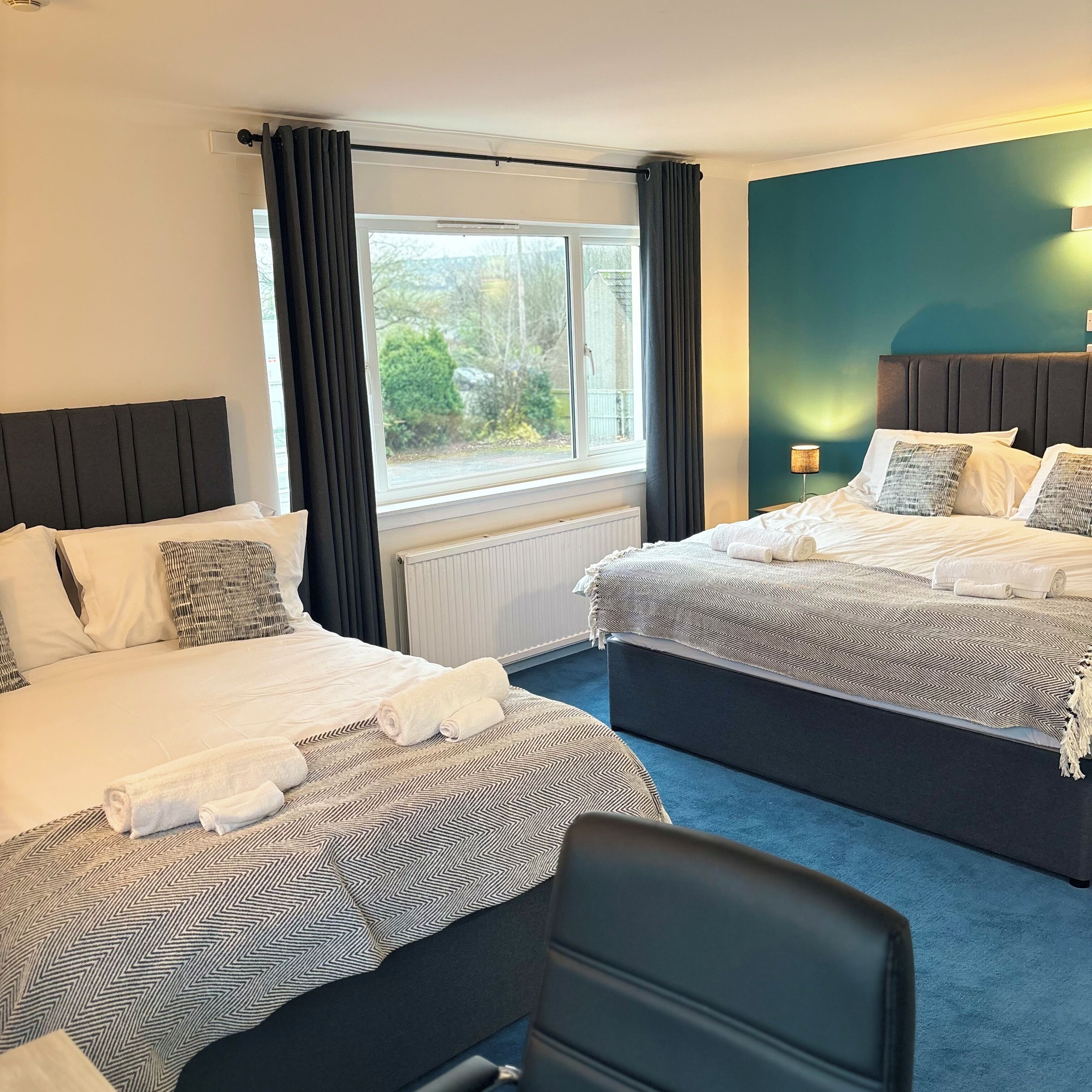 A hotel room with two beds, a view of greenery outside, and a blue carpet.
