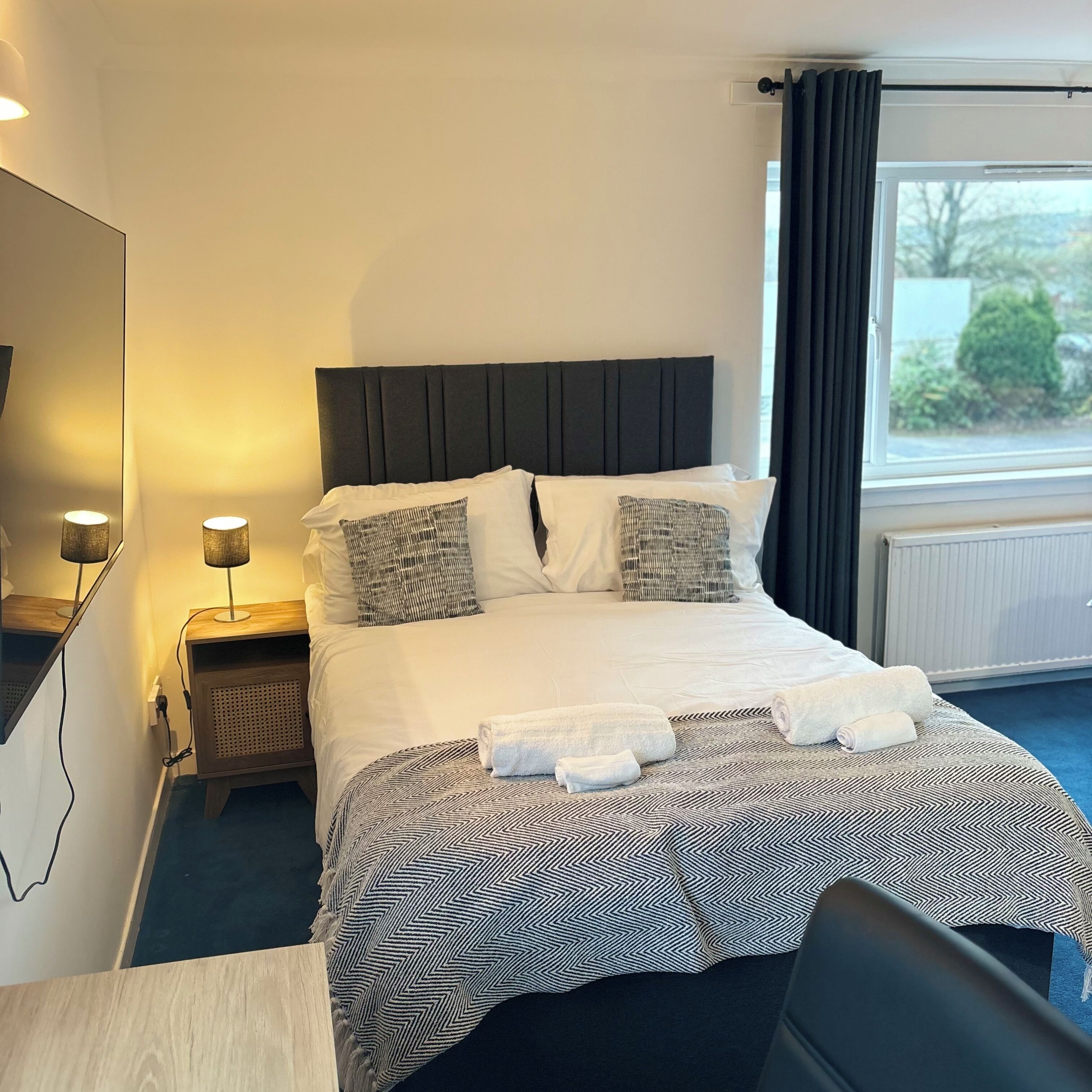 A hotel room with a bed, two bedside lamps, a window with curtains, and a view of greenery outside.