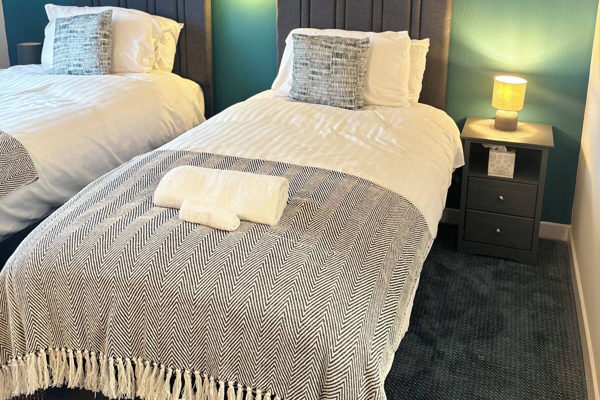 Ein Hotelzimmer mit zwei Betten, jedes mit einer anderen gemusterten Tagesdecke, einem Nachttisch mit Lampe und einer wandmontierten Leuchte.