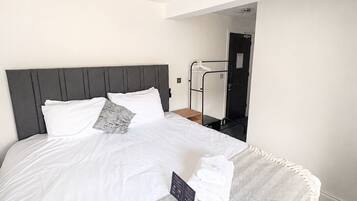 Habitación de hotel con cama, cabecera gris, cubrecama blanco, manta gris y blanca estampada, toalla y una pequeña señal negra sobre la cama.
