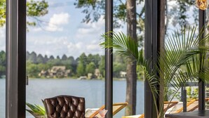 A modern room with large windows overlooking a lake, featuring a leather chair, wooden furniture, and potted plants. - NEW! 151 House on Lake Oconee (Eatonton)