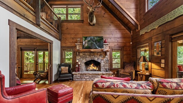 A log cabin interior with a stone fireplace, leather furniture, and a mounted deer head.
