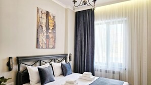 A neatly made bed with white and dark blue bedding, a chandelier, a large abstract painting, and a window with sheer curtains.