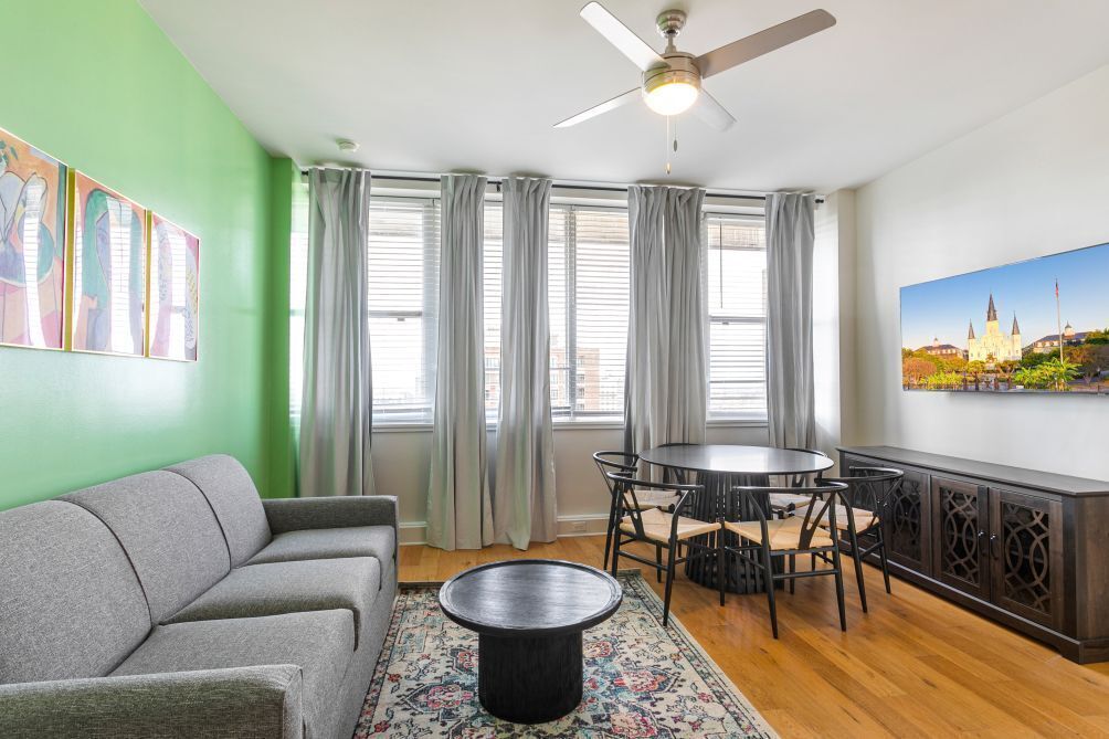Ein Wohnzimmer mit grauem Sofa, rundem Couchtisch, Esstisch mit Stühlen, Deckenventilator und großem Fenster mit Vorh ängen.