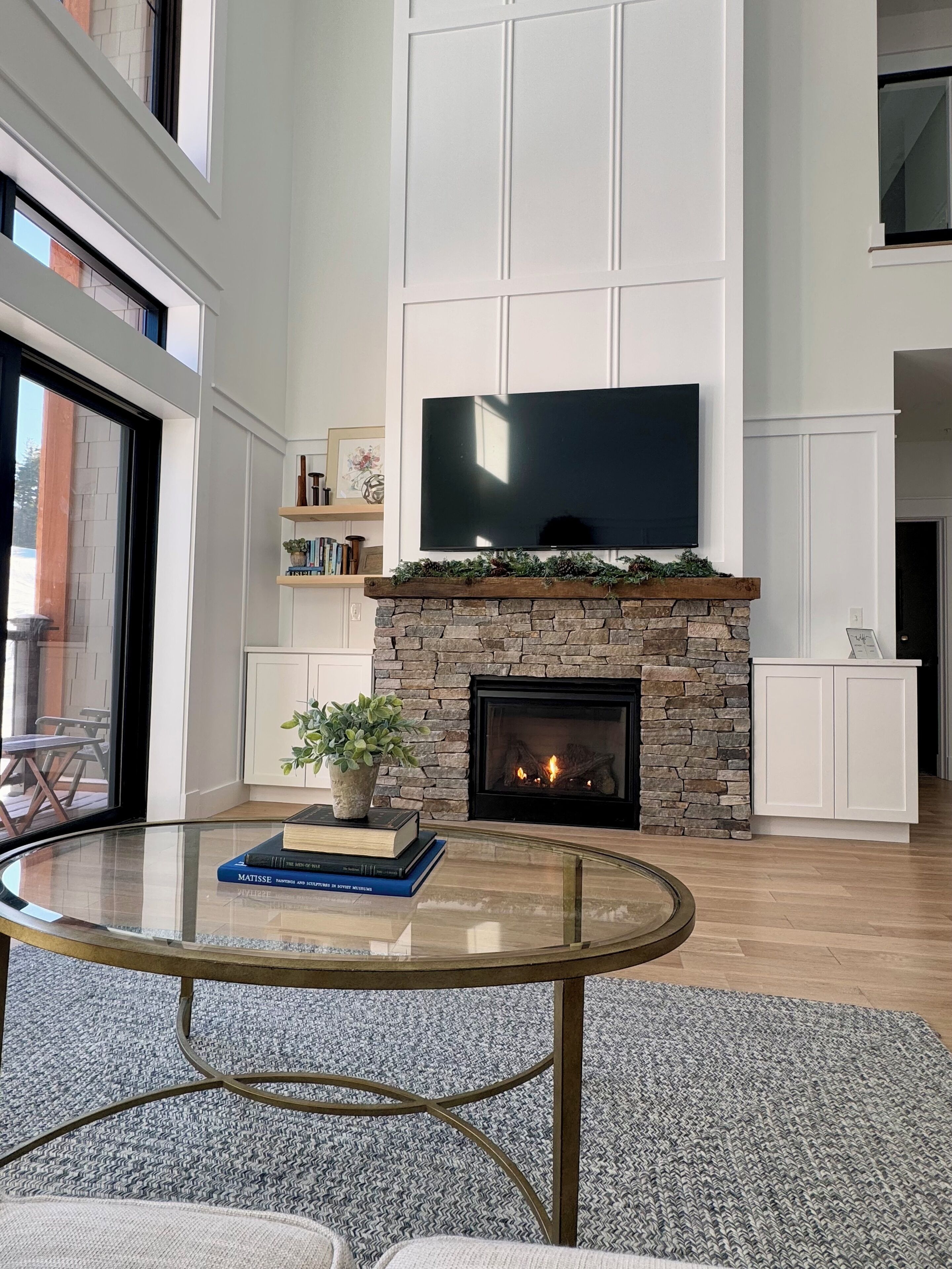A modern living room with a stone fireplace, a flat-screen TV mounted above it, a glass coffee table, and a bookshelf with books and decorative items.