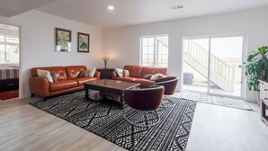 A modern living room with a brown leather sofa, a coffee table, and a patterned rug. There are two framed pictures on the wall, a staircase leading to a balcony, and a potted plant.