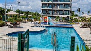 A pool area with a multi-level building in the background, surrounded by palm trees and outdoor seating. - Makia By Mai (Ipojuca)