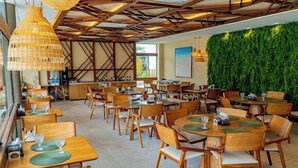 A restaurant with wooden tables and chairs, a green wall, and hanging wicker lamps. - Makia By Mai (Ipojuca)