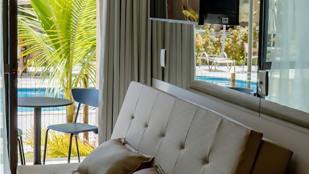 A modern living room with a sofa, a dining table, and chairs. There is a TV mounted on the wall and a window with curtains. - Makia By Mai (Ipojuca)