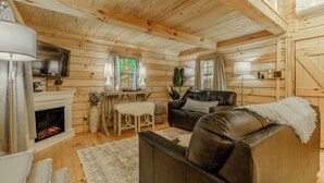 A log cabin interior with a fireplace, leather sofa, dining table, and wooden walls. - Sunset Serenity Luxury Cabin with Hot Tub Pet Friendly (Elizabethton)