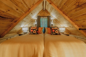 A room with a sloped ceiling, two beds with floral bedding, a window with curtains, and a wooden beam structure. - Monarch Mountain Luxury Cabin with Hot Tub Pet Friendly (Elizabethton)