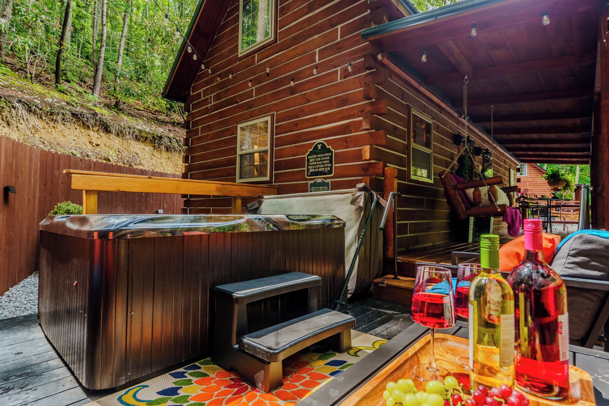 A wooden cabin with a hot tub, outdoor seating, and a wooden table with wine bottles and grapes.