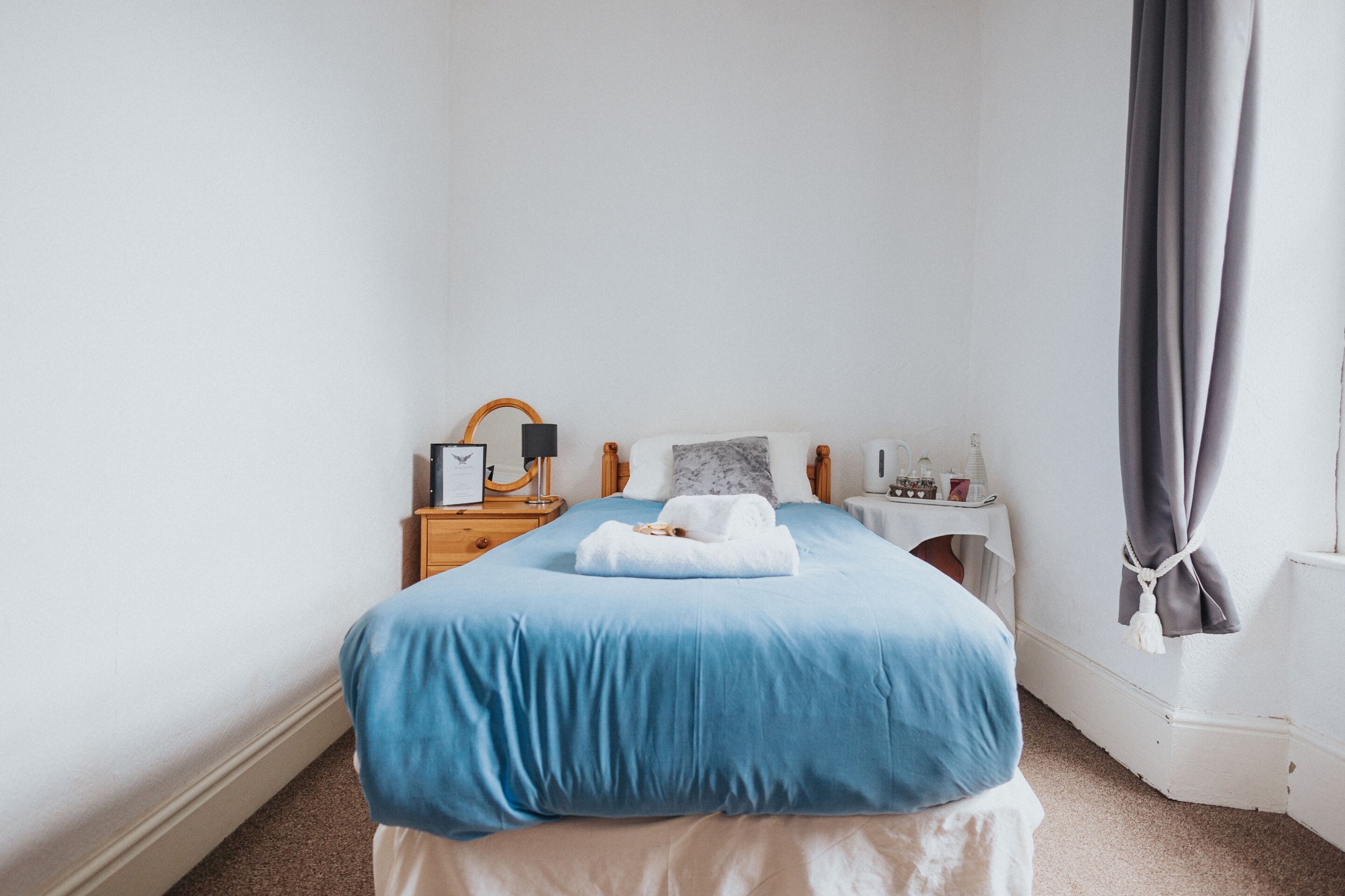 A neatly made bed with a blue bedspread and white pillows, a bedside table with a lamp, a small nightstand, and a window with curtains.