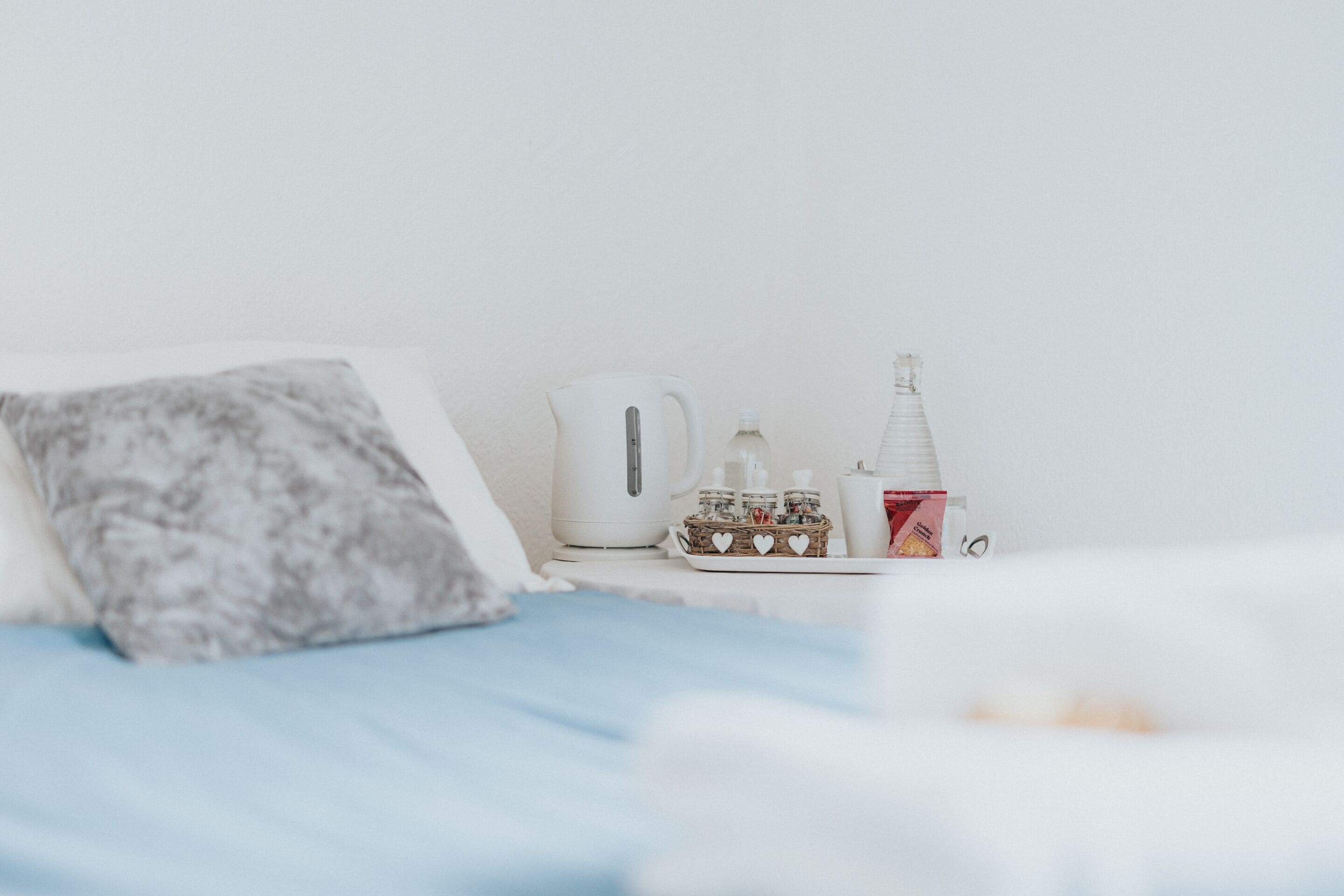 A neatly arranged hotel room with a bed, pillows, a kettle, a bottle, and a small basket of heart-shaped items.