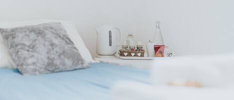 A neatly arranged hotel room with a bed, pillows, a kettle, a bottle, and a small basket of heart-shaped items.