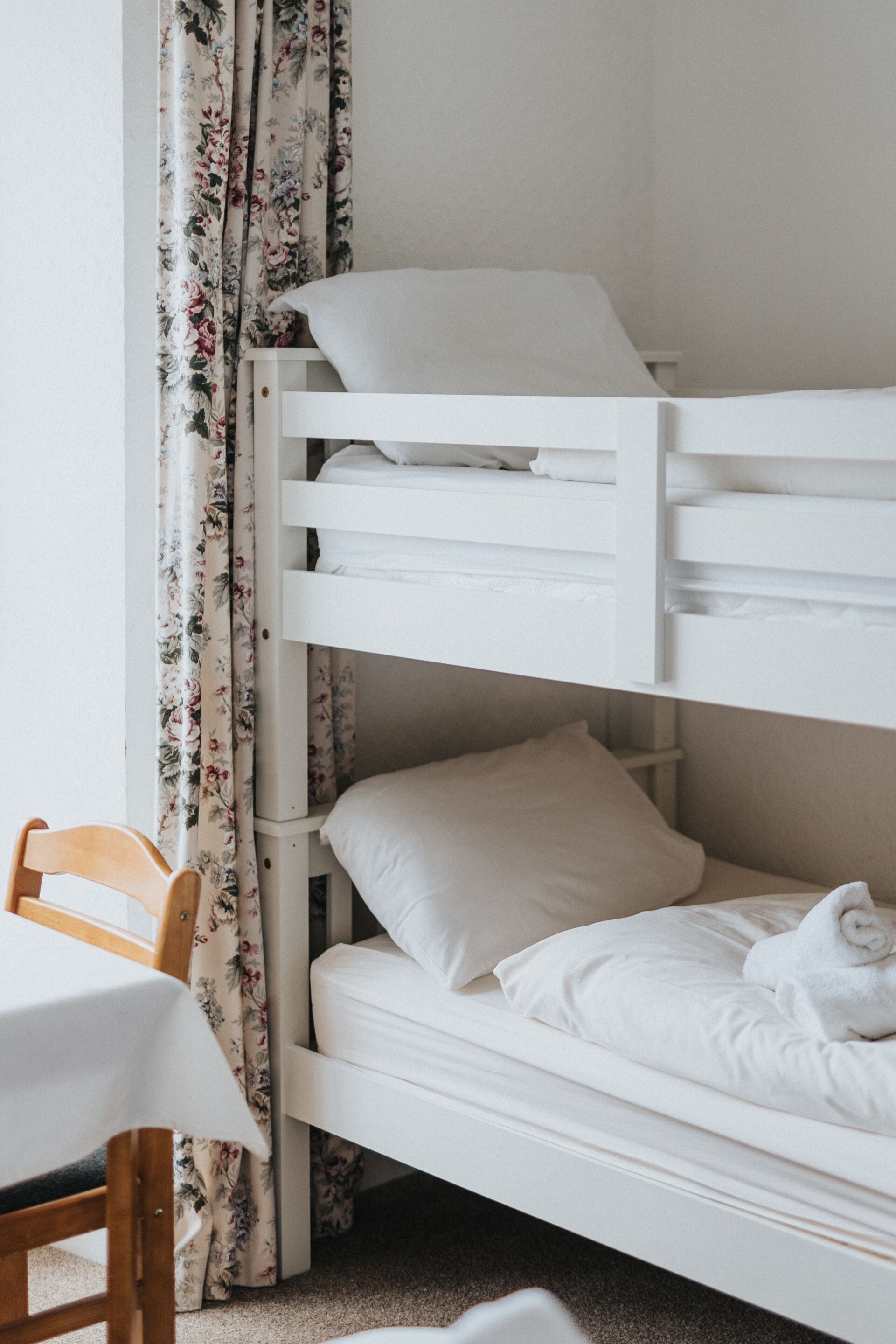 A bunk bed with white bedding, a wooden chair, and floral curtains.