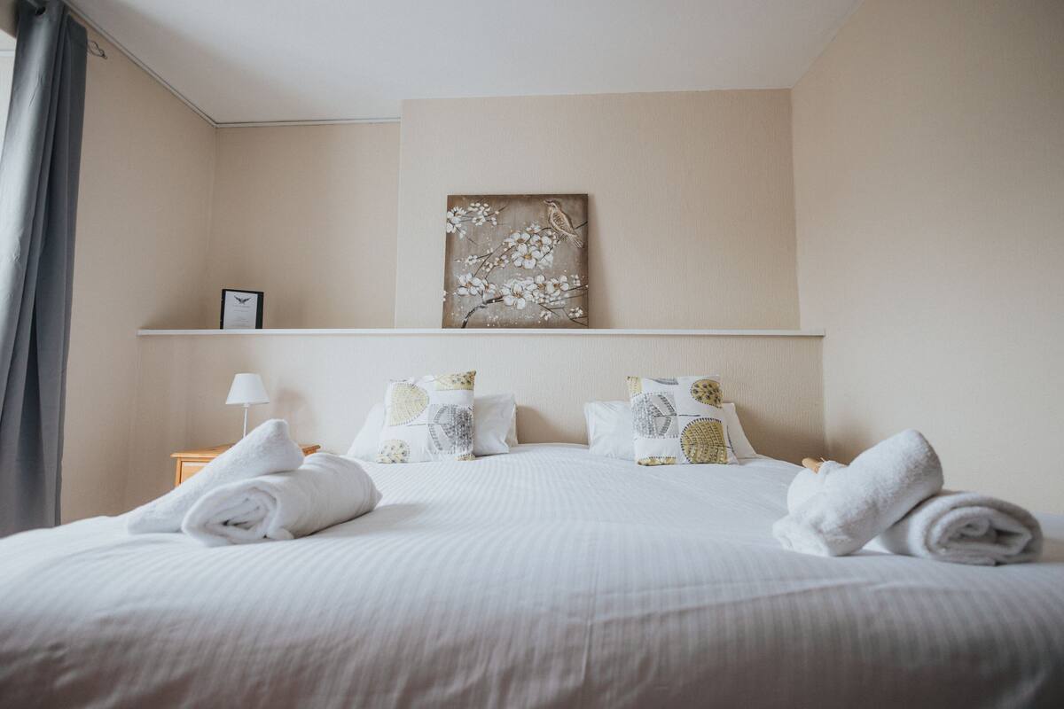 A neatly made bed with white linens and rolled towels, a wall-mounted picture of a bird on a branch, and a small bedside table with a lamp and a framed picture.