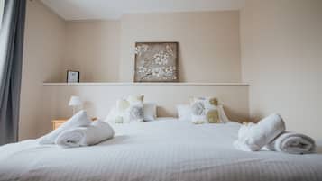 A neatly made bed with white linens and rolled towels, a wall-mounted picture of a bird on a branch, and a small bedside table with a lamp and a framed picture.