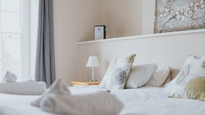 Een netjes opgemaakt bed met witte lakens en gestreepte kussens, een bedlampje en een ingelijst beeld aan de muur.