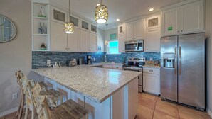 A kitchen with white cabinets, a granite countertop, a stainless steel refrigerator, and a blue-tiled backsplash. - Coastal Soiree Ocean Views, Game Room, & Steps From Port Aransas Beaches (Port Aransas)