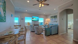 A spacious living room with a dining area, a ceiling fan, a TV, and a view of the ocean. - Coastal Soiree Ocean Views, Game Room, & Steps From Port Aransas Beaches (Port Aransas)