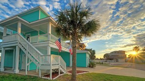 A two-story house with a balcony, a flag, and a palm tree in front. - Coastal Soiree Ocean Views, Game Room, & Steps From Port Aransas Beaches (Port Aransas)