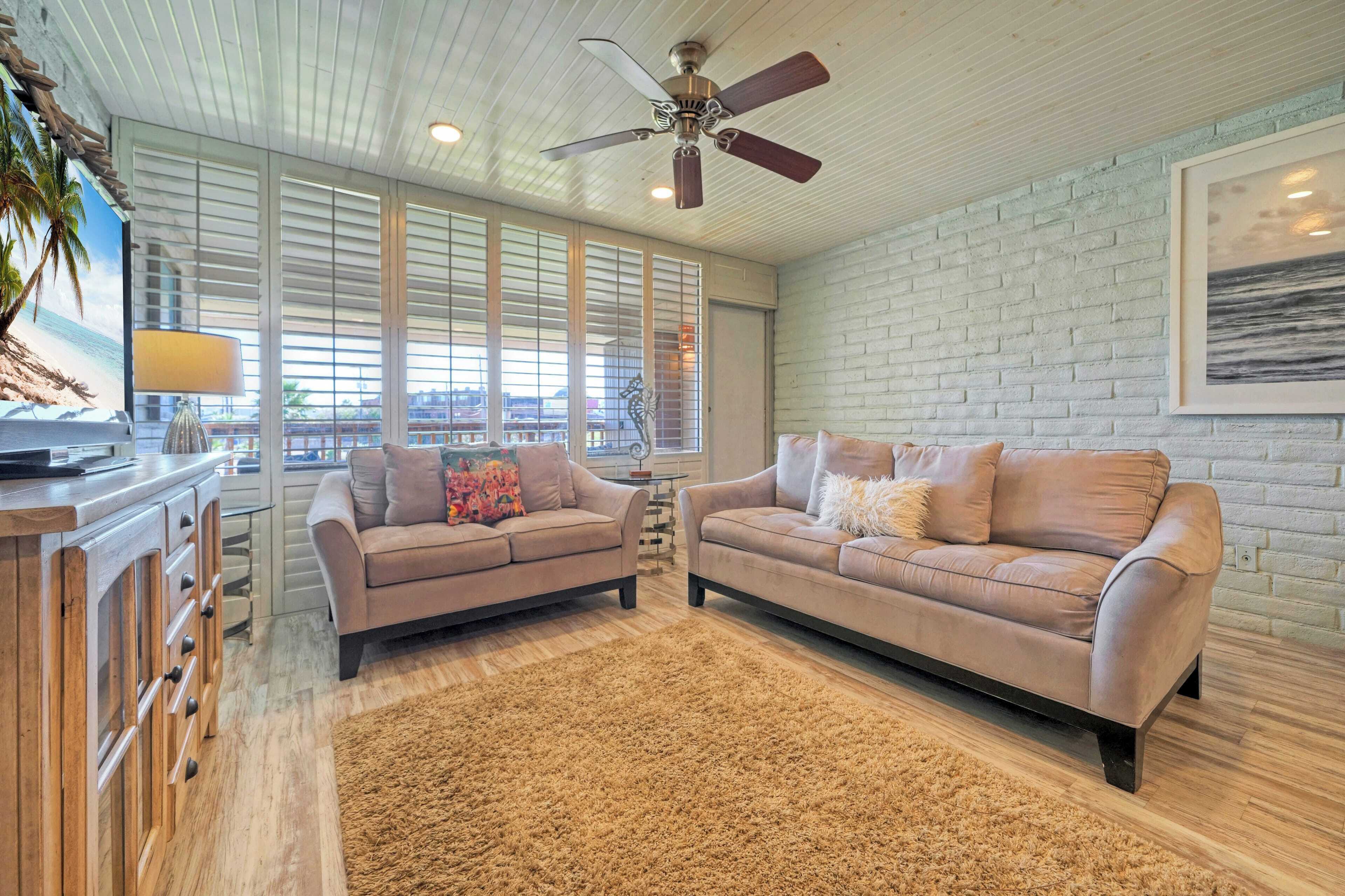 A living room with two sofas, a ceiling fan, large windows, and a view of the beach.
