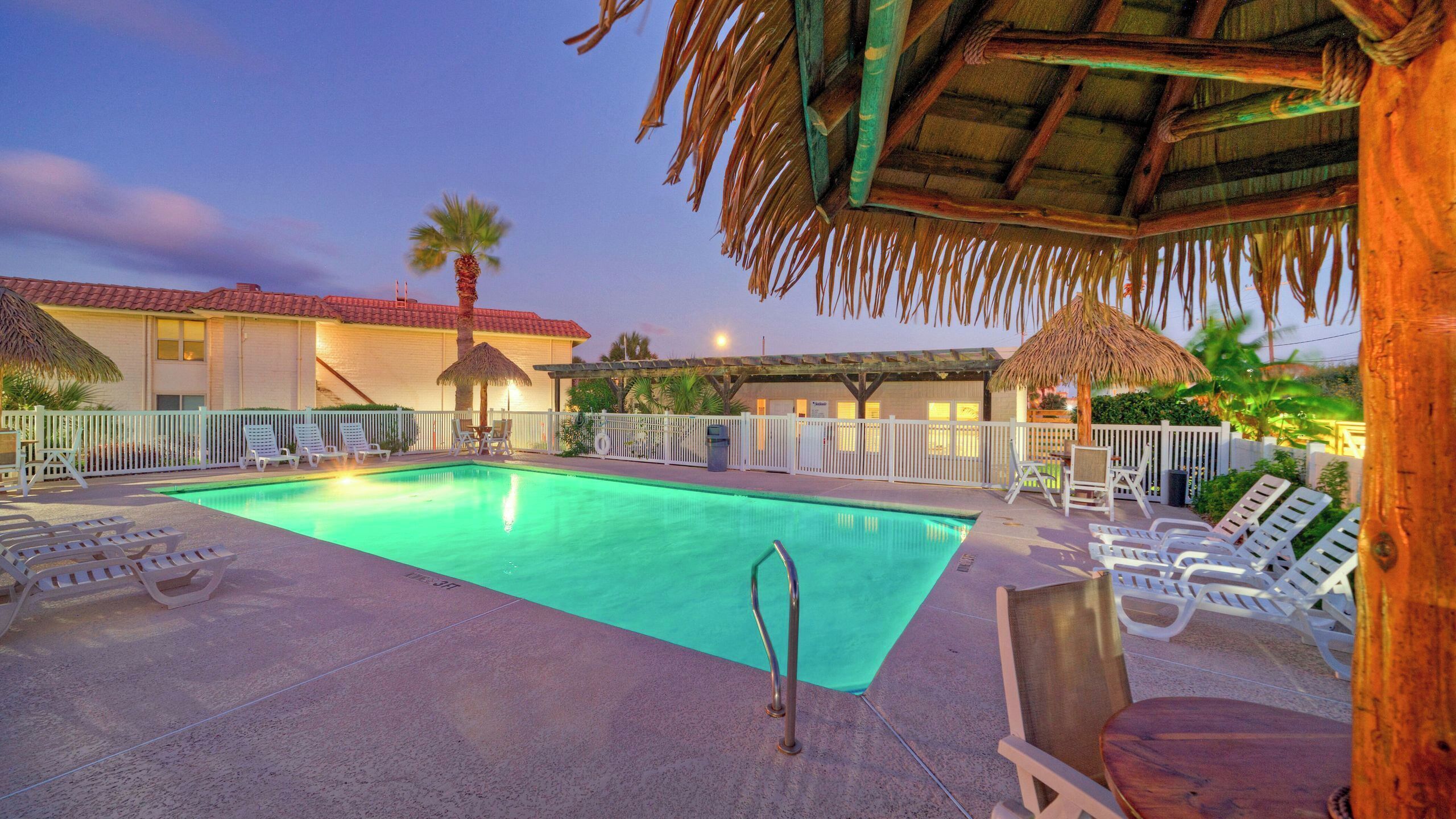 A swimming pool with lounge chairs, a thatched roof, and a fenced area.